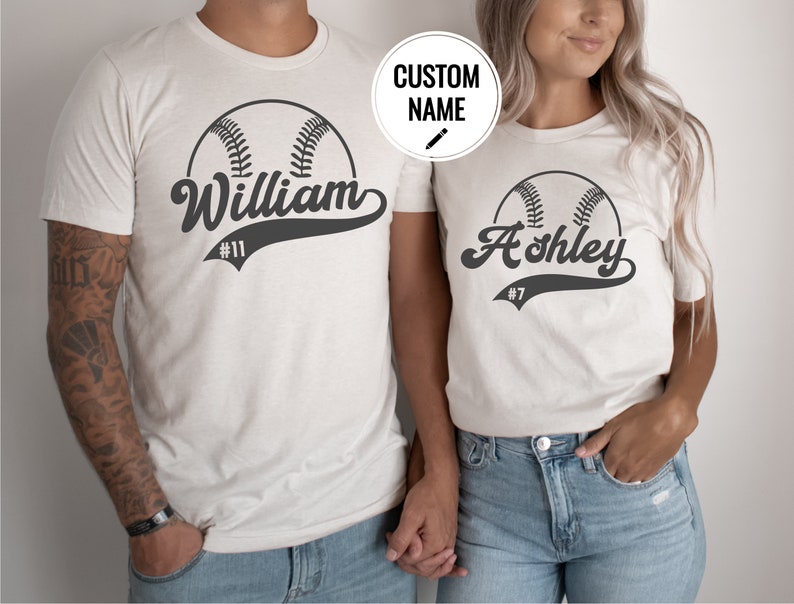 May include: Two white t-shirts with a baseball graphic and personalized names. The shirt on the left says "William #11" and the shirt on the right says "Ashley #7".