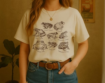 Könnte beinhalten: Ein cremefarbenes Kurzarm-T-Shirt mit einem dunkelbraunen Grafikdruck verschiedener Froscharten, darunter der Nördliche Leopardfrosch, Ochsenfrosch, Graulaubfrosch, Rotaugenlaubfrosch, Panama-Stummelfußfrosch, Tomatenfrosch und Schmuckhornfrosch.