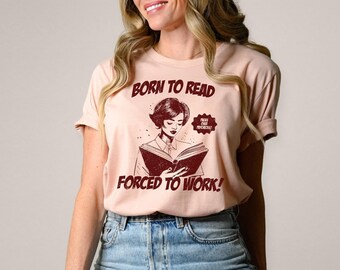 May include: A light pink short-sleeve t-shirt featuring a dark red graphic of a person reading a book. The text reads BORN TO READ FORCED TO WORK! and a smaller speech bubble says PAGES OVER PAYCHECKS! The shirt has a crew neckline and rolled sleeves.