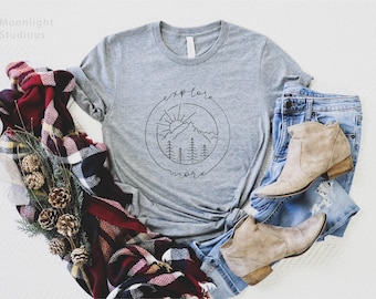 May include: A heather gray short-sleeve t-shirt featuring a black circular line art graphic of mountains, pine trees, and a sun with the text "explore more". The shirt is paired with blue denim jeans, tan suede ankle boots, and a plaid scarf.