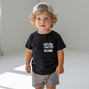 A young child with curly blonde hair wearing a black t-shirt with a graphic of a race car and the name "OLIVER" printed on it, along with a baseball cap.