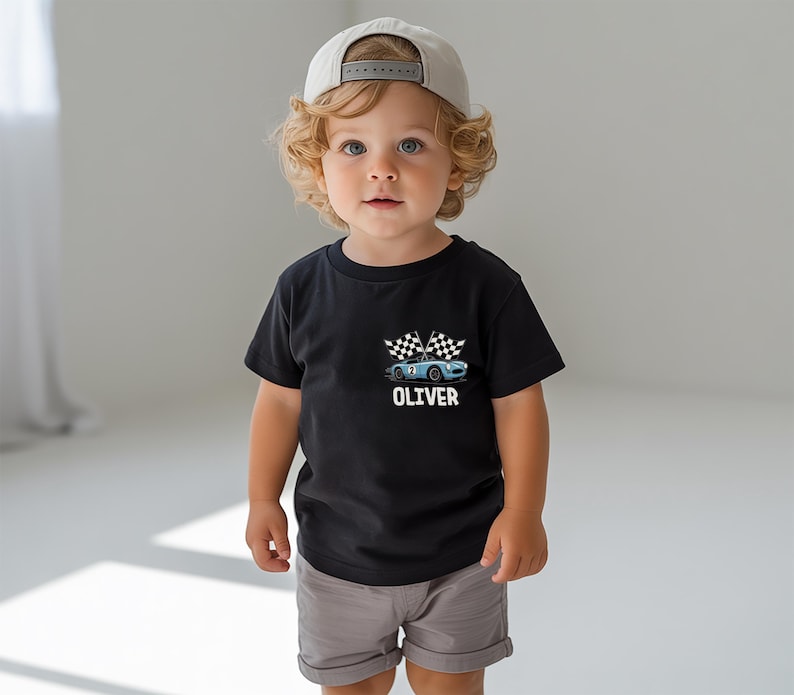 A young child with curly blonde hair wearing a black t-shirt with a graphic of a race car and the name "OLIVER" printed on it, along with a baseball cap.