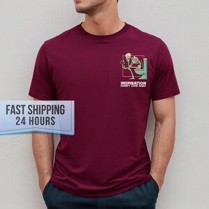 May include: A maroon short-sleeved t-shirt featuring a graphic on the left chest. The graphic depicts a skeleton sitting on a green toilet, holding a lit match, with the text "INSPIRATION DOESN'T COME EASILY" printed in white capital letters below it.