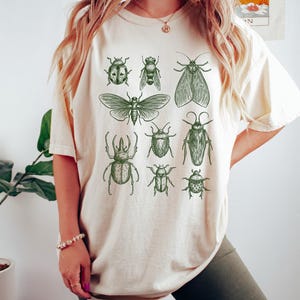 May include: A cream-colored short-sleeve t-shirt featuring a dark green graphic print of various insects, including beetles, a moth, and a cicada. The design is arranged in a grid-like pattern across the center of the chest area of the shirt.