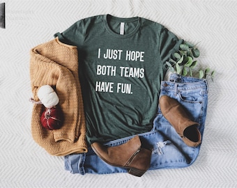 以下が含まれることがあります： 白い文字で「I JUST HOPE BOTH TEAMS HAVE FUN」と書かれたダークグリーンの半袖Tシャツ。マスタードイエローのニットセーター、ブルーのデニムジーンズ、ブラウンのアンクルブーツ、そして小さな赤と白の装飾品が一緒に配置されています。