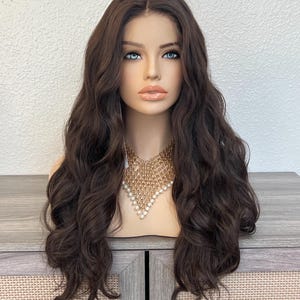 May include: A brown wig with long, wavy hair is displayed on a mannequin head. The wig features a natural-looking hairline and is styled with loose curls. The mannequin is wearing a gold-toned necklace with pearl accents.