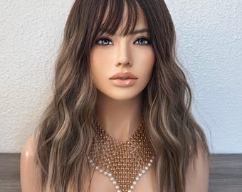 May include: A shoulder-length wig with wavy brown hair, featuring a darker root color and lighter highlights throughout. The hair is styled with straight-cut bangs that fall across the forehead. A gold-toned mesh necklace with white pearls.