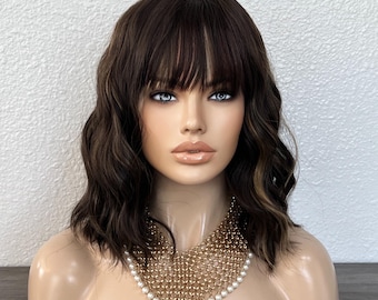 May include: A brown wig with bangs and wavy layers is displayed on a mannequin head. The wig has highlights and is styled to frame the face. A gold-toned necklace with pearl accents is draped around the mannequin's neck.
