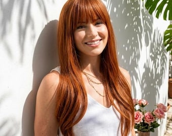 May include: A woman with long, auburn hair and bangs smiles, wearing a white tank top and a layered gold necklace. The background features a white wall with palm shadows, a vase of pink roses, and green tropical leaves.