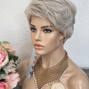 May include: A mannequin head with a short, light gray wig and a gold-tone chain necklace with faux pearls. The necklace has a circular pendant with the word "lum" on it. A tag with a pattern is visible.