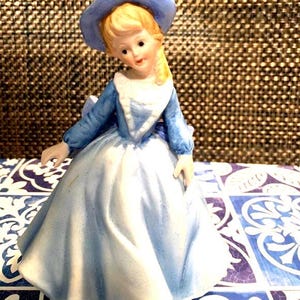 Puede incluir: Una figurita de porcelana de una niña pequeña con un vestido y un sombrero azules. Tiene el pelo rubio y los ojos azules. La figurita está sentada sobre una superficie de azulejos.