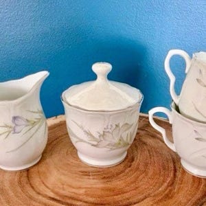 May include: A white porcelain sugar bowl with a lid and a floral design, along with two matching creamer and teacups.