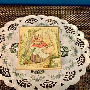 Puede incluir: Una caja de madera rectangular con una ilustración de un conejo con capa roja y cesta, en una escena de bosque. La caja está sobre un mantel individual blanco con patrones florales y de hojas. El fondo es una superficie tejida marrón oscuro.