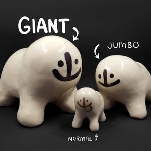 Puede incluir: Tres figuras de cerámica blanca con forma de foca de diferentes tamaños, cada una con una cara sonriente negra. La más grande está etiquetada como "GIANT", la mediana como "JUMBO" y la más pequeña como "NORMAL". Las figuras están sobre un fondo negro.