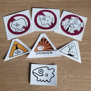 Puede incluir: Conjunto de diez pegatinas con diversas ilustraciones en blanco y negro de peces de dibujos animados, una criatura con una corona y una señal de advertencia que dice "DANGER". Las pegatinas están sobre una superficie de madera marrón claro.