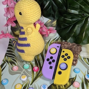 May include: A purple and yellow Nintendo Switch controller with custom leaf-shaped buttons. The controller is surrounded by small, colorful leaf-shaped buttons and a yellow crocheted dinosaur toy.
