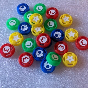May include: A collection of colorful round buttons with various Nintendo characters and symbols, including Mario, Luigi, stars, and question marks. The buttons are made of silicone and are designed to be used as replacements for the standard buttons on a Nintendo Switch controller.