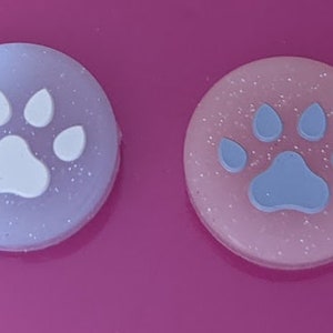 May include: Two round, glittery paw print buttons. One button is light blue with a white paw print. The other button is pink with a blue paw print.