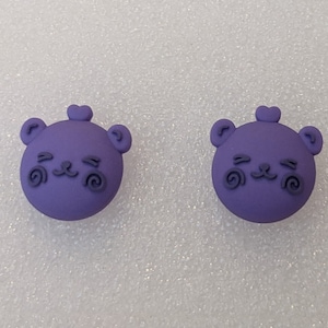 May include: A pair of purple bear-shaped earrings with a smiling face and swirl details.