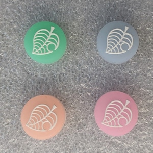 May include: Four round, silicone button covers for a video game controller. The covers are in four different colors: light green, light blue, light pink, and peach. Each cover has a white leaf design with a small circle in the center.