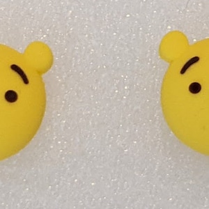 May include: Two yellow Winnie the Pooh character face earrings. The earrings are round and have a black outline of Winnie the Pooh's face with black eyes and a black nose.