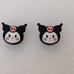 May include: Two black and pink rubber charms featuring a cartoon character with a skull on its head. The character has a pink face with black eyes and a pink mouth.