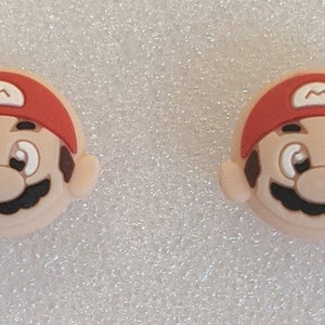 May include: Two red and brown cartoon character face charms with a white M on the red hat. The character has a brown mustache and eyebrows, a brown nose, and white eyes.