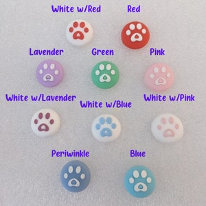 May include: Assortment of ten round, paw print-shaped buttons in various colors, including white, red, lavender, green, pink, blue, and periwinkle. Each button has a white paw print with a heart in the center.