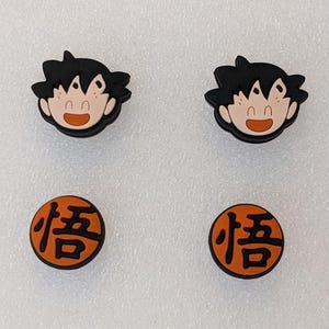 May include: Four shoe charms featuring characters from Dragon Ball Z. Two charms depict Goku's face with black hair and an orange mouth. The other two charms are orange circles with black Japanese characters.