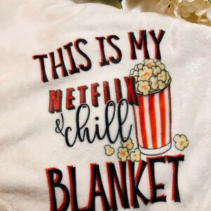 K&ouml;nnte beinhalten: Eine wei&szlig;e Pl&uuml;schdecke mit dem Text "This is my Netflix & chill blanket" in schwarzer und roter Schrift. Neben dem Text ist ein rot-wei&szlig; gestreifter Eimer mit Popcorn abgebildet.