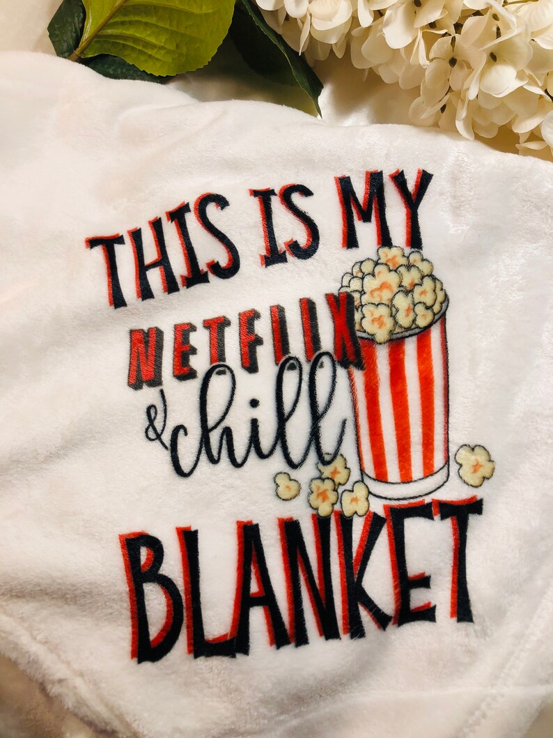 K&ouml;nnte beinhalten: Eine wei&szlig;e Pl&uuml;schdecke mit dem Text "This is my Netflix & chill blanket" in schwarzer und roter Schrift. Neben dem Text ist ein rot-wei&szlig; gestreifter Eimer mit Popcorn abgebildet.