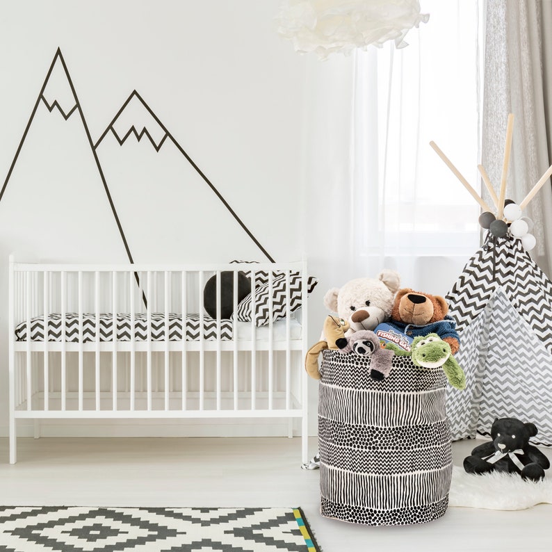 May include: A black and white patterned fabric storage bin filled with stuffed animals, including a teddy bear, a monkey, and a frog. The bin is sitting on a white floor in a nursery with a white crib and a teepee tent in the background.