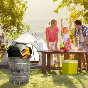 May include: A family of three enjoys a camping trip, preparing food on a wooden table. A black and white striped canvas storage bin sits in the foreground, filled with camping gear. A white tent is in the background.