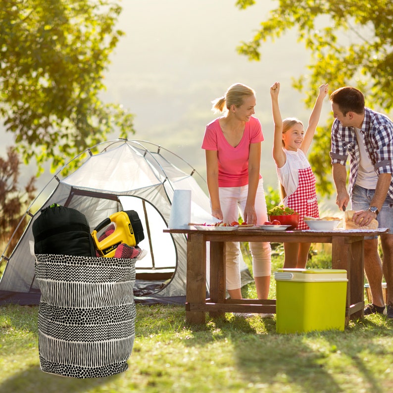 May include: A family of three enjoys a camping trip, preparing food on a wooden table. A black and white striped canvas storage bin sits in the foreground, filled with camping gear. A white tent is in the background.