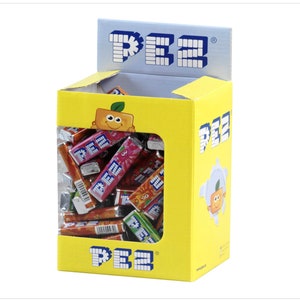 May include: A yellow box filled with assorted PEZ candy sticks. The box has a clear window showing the candy. The box has a PEZ logo on the front and a cartoon orange with a smiling face.