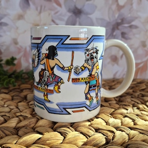 May include: White ceramic mug featuring colorful illustrations of Native American Kachina figures. The mug has a curved handle and a blue and white geometric border design. The words "HON KACHINA" and "OWIK KACHINA" are printed on the mug.