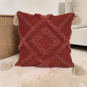 May include: A square, terracotta-coloured cushion featuring a textured diamond pattern and four cream-coloured tassels. The cushion has a woven texture and is placed on a white surface.