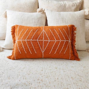 May include: A rectangular burnt orange throw pillow featuring a white embroidered geometric diamond pattern across the center. The pillow has textured fabric and matching orange fringe trim along the two shorter side edges of the rectangular cushion.