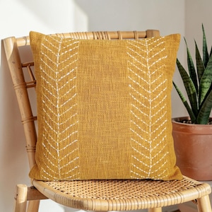 May include: Mustard yellow square throw pillow with white embroidered leaf pattern. The pillow is made of textured fabric and is placed on a woven bamboo chair. A potted plant is visible in the background.