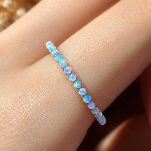 Op de afbeelding: Een dunne, verfijnde ring met een doorlopende rij kleine, ronde, iriserende opalen. Elke opaal vertoont flitsen van blauw, roze en groen licht, gezet in een slanke, gepolijste zilverkleurige metalen band die om de vinger zit.