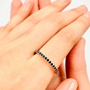 Può includere: Un anello d'argento con una fila di piccole pietre nere rotonde. L'anello è indossato su un dito, tenuto da un'altra mano. Il gioiello è un design delicato.