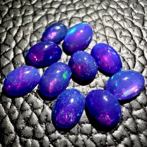 May include: A collection of oval-shaped opals in shades of deep blue and purple, with flashes of iridescent color. The gemstones are arranged on a textured black surface, creating a striking contrast.