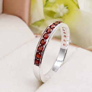 Op de afbeelding: Een zilveren ring met een kanaalzetting met een rij kleine, ronde, dieprode granaatedelstenen. De ring staat rechtop in een lichtgekleurd, gevoerd juwelendoosje, waarbij de metalen glans en de donkerrode stenen duidelijk zichtbaar zijn.