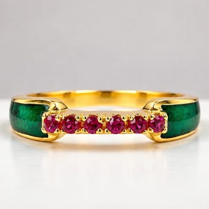 Puede incluir: Un anillo de oro con una fila central de seis pequeñas piedras preciosas redondas de color rojo rosado vibrante, flanqueadas por dos incrustaciones de esmalte verde intenso a cada lado de la banda, creando un contraste entre el metal, la piedra y el esmalte.