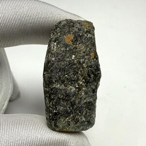 Könnte beinhalten: Ein raues, dunkelgraues Mineralexemplar mit strukturierter Oberfläche und kleinen, hellbraunen Flecken. Der Stein wird vor einem hellgrauen Stoffhintergrund gehalten, der sein natürliches, unpoliertes Aussehen zeigt. Die Form ist länglich und unregelmäßig.