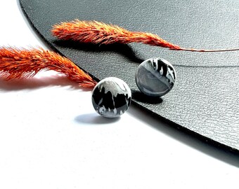 May include: Two round, gray and black decorative beads with a marbled design. The beads are placed on a textured black surface, with a dried orange plant stem in the background. The beads are a unique accessory.
