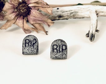 May include: Two gray tombstone-shaped earrings with the letters "RIP" in black. The earrings are covered in silver glitter. The background includes a dried flower and a piece of driftwood, suggesting a Halloween or gothic theme.