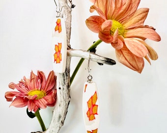 May include: A pair of rectangular earrings with a white background and a colorful design of orange and yellow shapes. The earrings are hanging from silver hooks. The image also features a piece of driftwood and two peach-colored flowers.