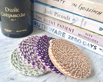 May include: A set of four circular coasters made from braided rope in shades of purple, tan, and light green. The coasters are stacked in a staggered row, featuring a spiral pattern created by the woven cord construction with a flat, round shape.