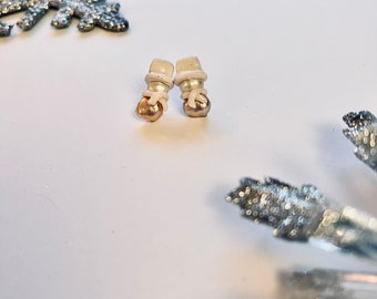 May include: A pair of snowman-shaped earrings. Each earring features a white hat, a beige body, and a gold-colored base. The earrings are set against a white background with silver snowflake decorations.